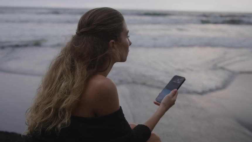 Close up of a woman browsing her smartphone sitting next to the shore of a beach at dusk