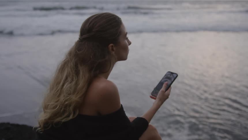 Woman sitting on the beach using a smartphone with the ocean in the background