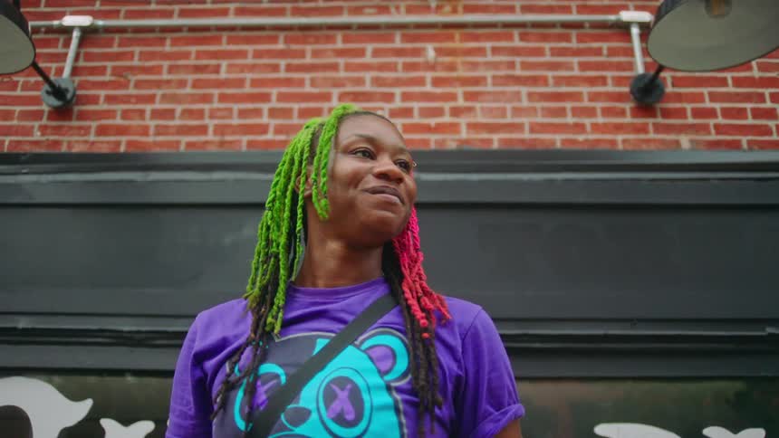 A woman with a purple tshirt and multicolored braided hair stands in front of a brick wall