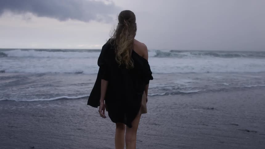 A woman wearing a black dress walking on a beach facing the ocean