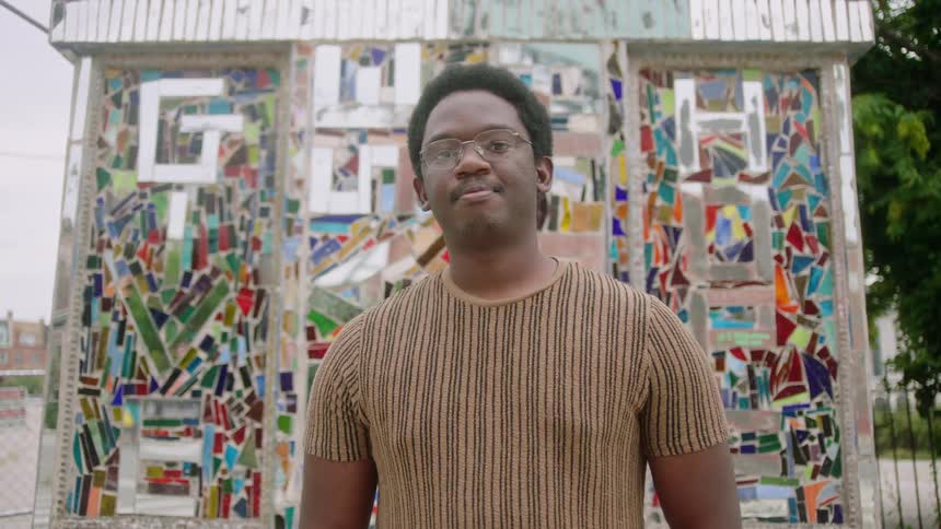 A black man in glasses and a brown shirt is standing in front of a public art work made of stained glass the words give and hope are visible in the artwork behind him