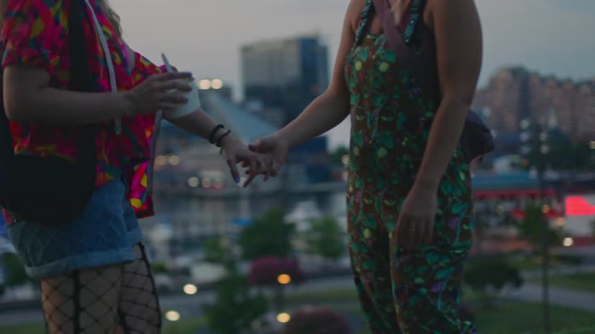 Two women holding hands wearing fashionable and colorful clothes walking in the late afternoon with buildings in the background of a metropolis