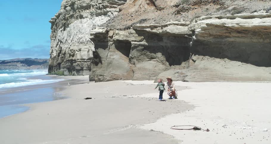 Mother on the beach sand with her little daughter next to the rocky coast