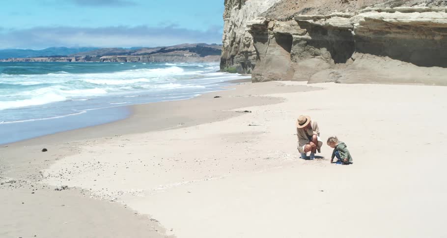 A mother and her daughter crouch on a sandy beach with waves rolling in from the sea next to a cliff on a sunny day