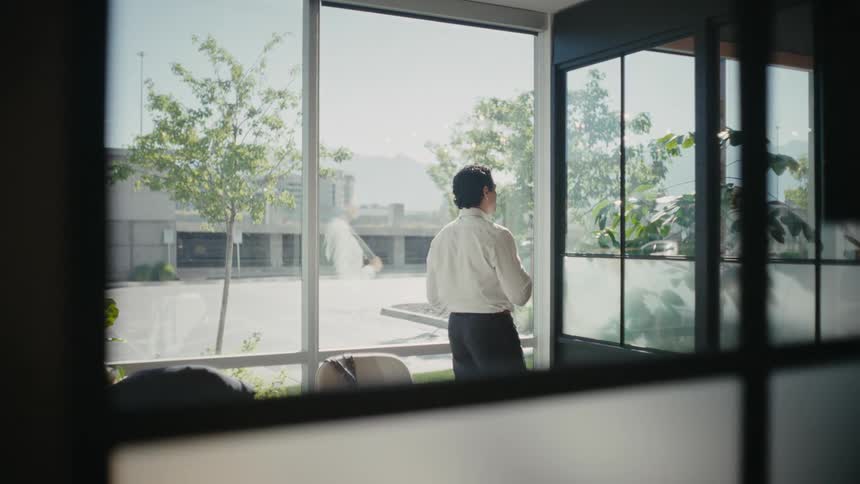 A man wearing a business attire stands on an office with windows looking at a city street