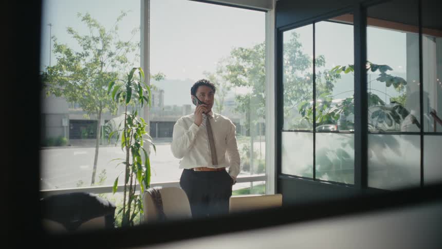 A man in a white shirt standing by a window in a welllit room on a phone call