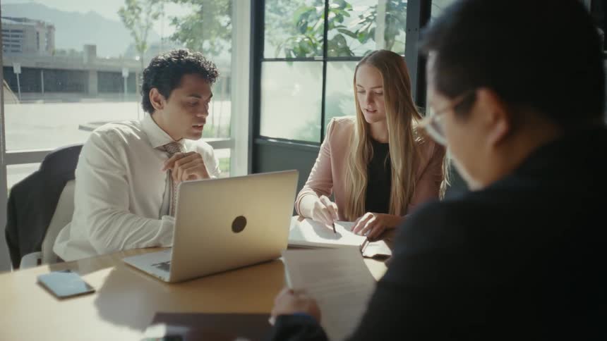 Three people sitting at an office table with documents and a laptop talking