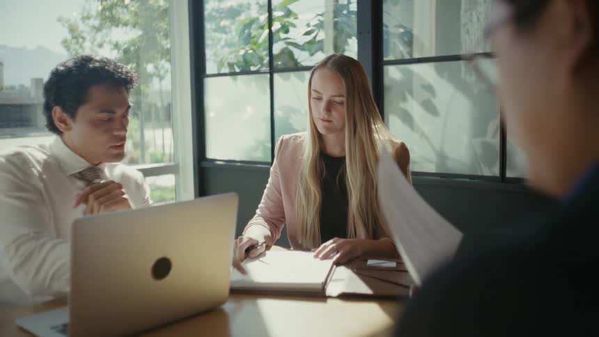 Three people sitting at an office desk talking and with documents and laptop on the table