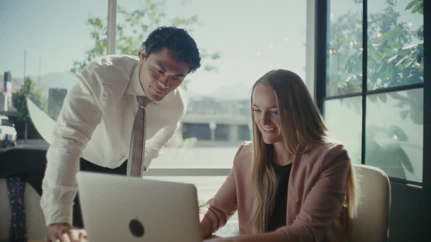 Man and a woman at an office desk talking and with a laptop in front