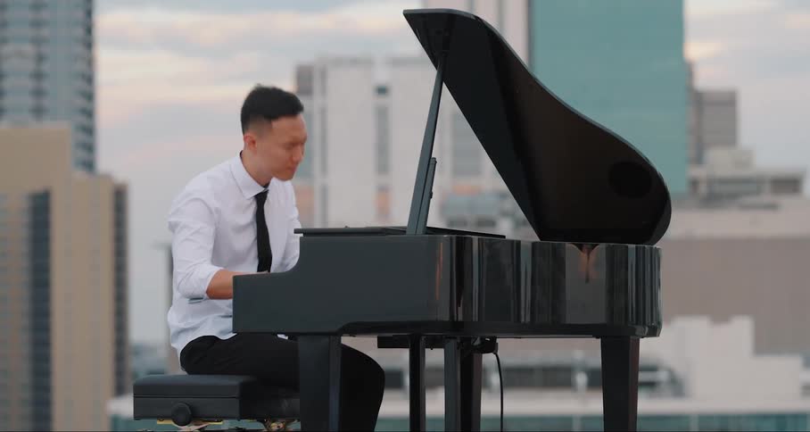 A musician seated at a grand piano on a highrise buildings rooftop with the city skyline in the background