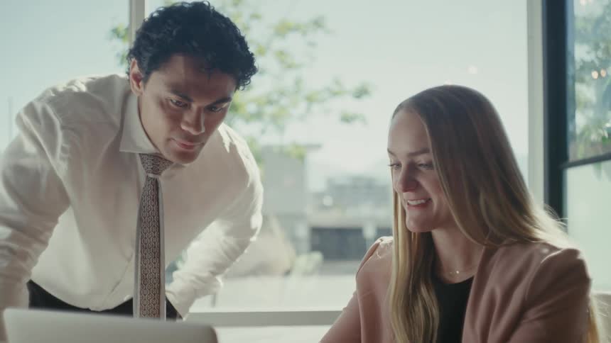 A professional office setting with coworkers reviewing work on a laptop next to a large window