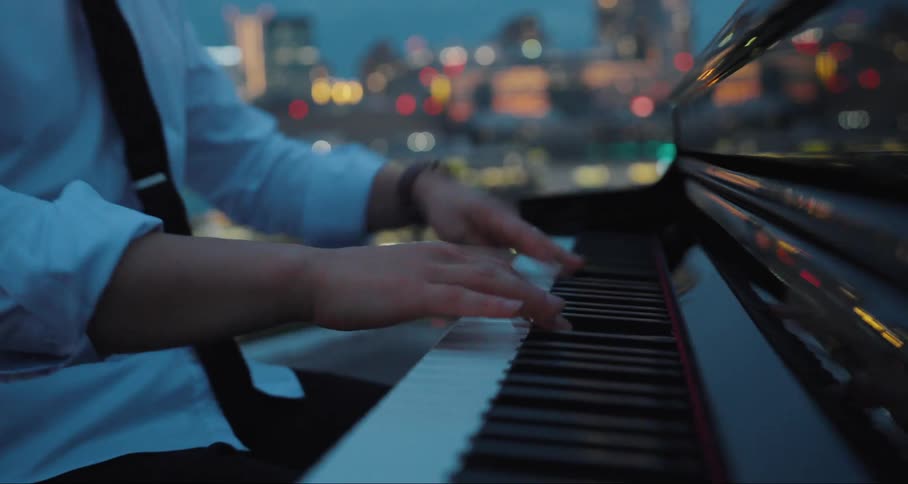 A closeup of hands playing a piano against a blurred cityscape during twilight hours