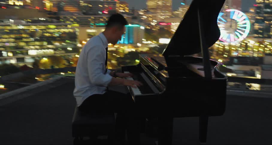 Man playing piano on terrace with buildings lit up at night around