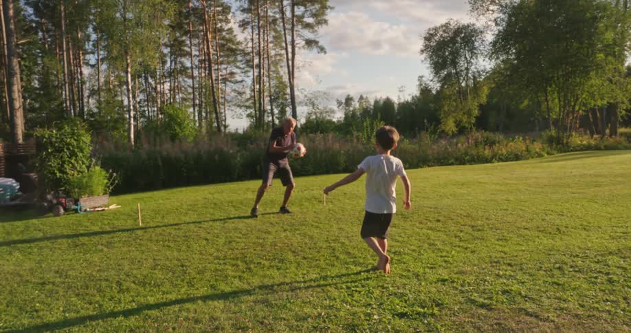 Boy kicking the ball to his grandfather who catches it with his hands and returns it while they are in the backyard of a house surrounded by vegetation