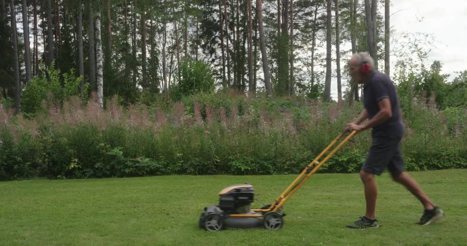 An elderly man mows grass near a tree grove on a cloudy day