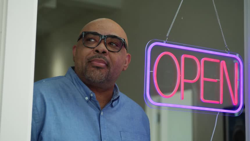 A man turns on the open sign hanging in the window and pauses smiling