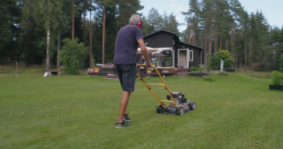An elderly man mowing the lawn with a yellow and black lawnmower in front of a red house