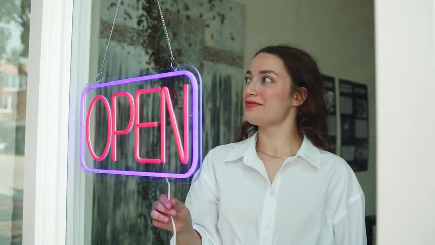 A woman stands behind a glass door turning on a neon sign reading open in bright pink and blue colors