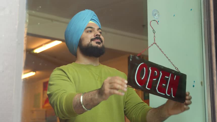 Man with a blue cloth on his head turning a sign in a store window with the signage written open