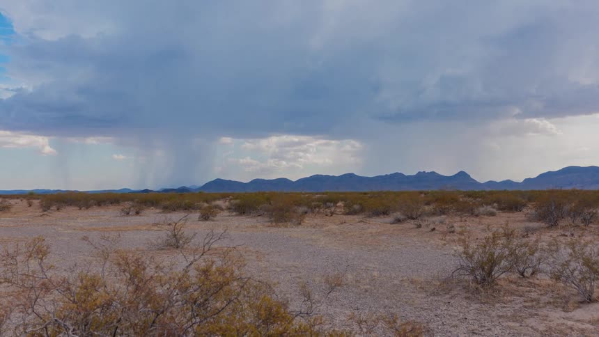 A desert landscape under a cloudy sky rain falling in the distance next to a mountain range