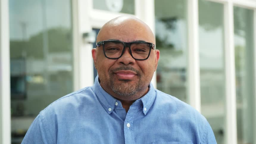 A man with glasses and a short beard wearing a light blue button up shirt standing outdoors in front of a building with large windows