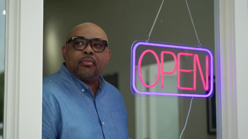 A man stands behind a glass door turning off a neon sign reading open in bright pink and blue colors