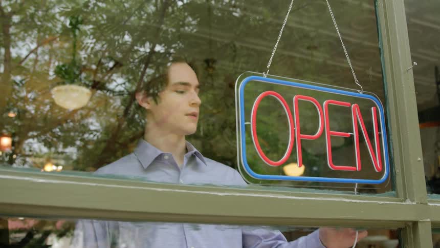 A young man turns on a neon sign reading open in bright pink and blue colors hanging inside a window
