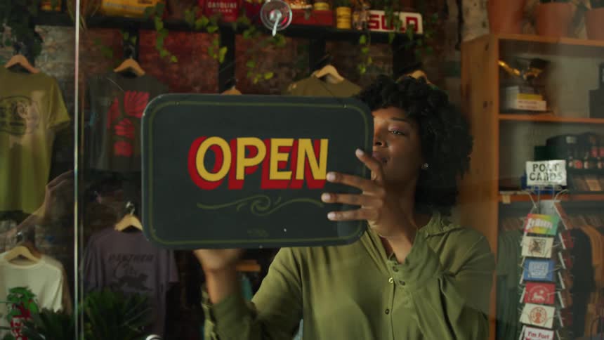 A woman stands behind a glass door turning around a sign reading open in bright red and yellow colors