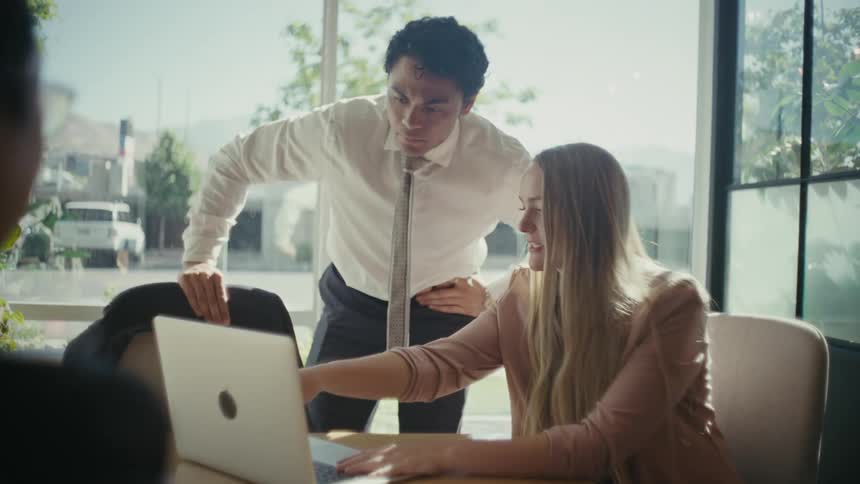 A professional office setting with coworkers reviewing work on a laptop next to a large window