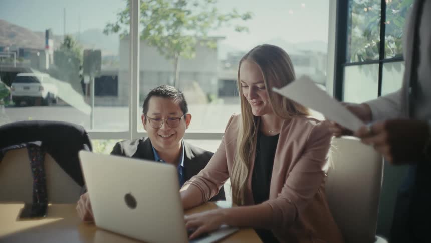 A modern office scene with one person seated at a table working on a laptop while another crouches next to them
