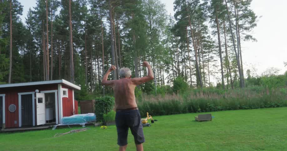 Elderly man with his back turned shirtless with his arms raised up and in the background his little grandson as they stand in a yard with grass and trees around