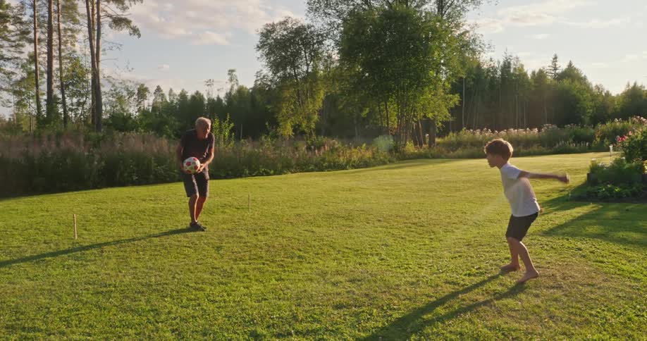 Boy practices kicking a soccer ball with the help of his grandfather on the lawn on a cloudy summer day