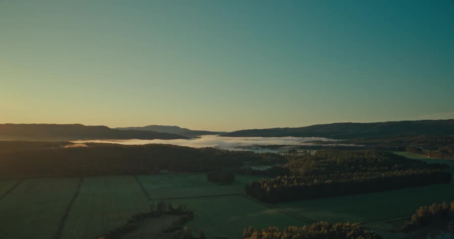An aerial view of a rural landscape at sunrise or sunset with light fog over fields patches of distant forest and gentle hills on the horizon