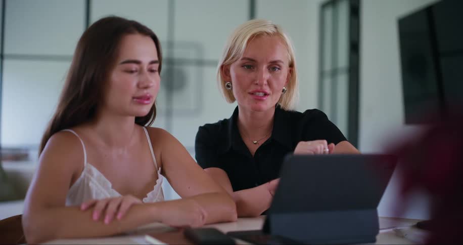 Two women sitting at a table facing a laptop one person is gesturing towards the screen explaining something