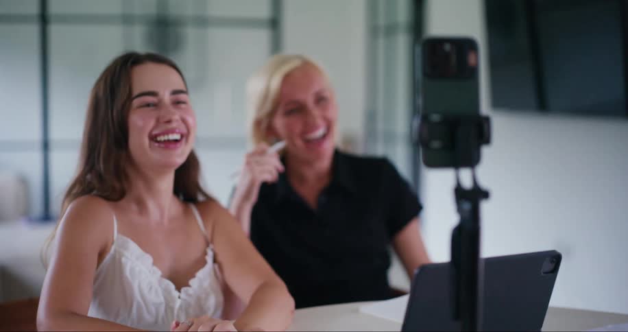 Two friends sit and laugh while an iphone is in selfie mode in front of them