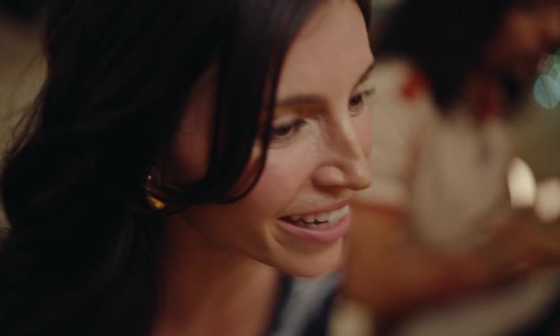 A smiling woman with dark hair engaged in conversation while a blurred figure is visible in the background