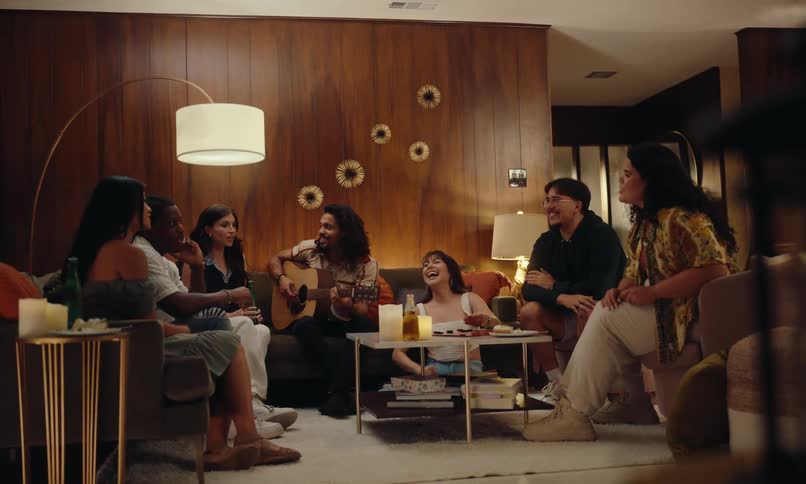 Group of friends sitting in a living room at night talking and laughing with one of them holding a guitar