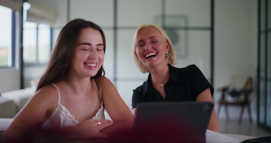 Two young women sitting side by side laughing in front of a tablet