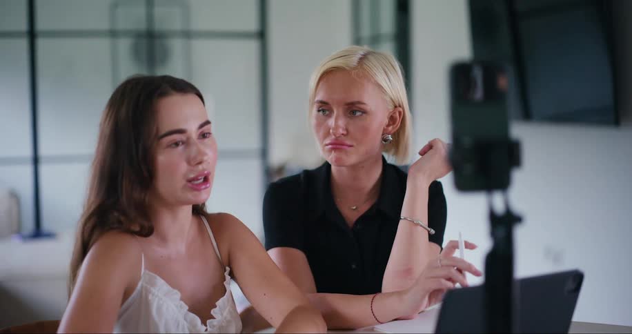 Two women are talking and speaking to a cell phone that is in front of them attached to a tripod and filming them