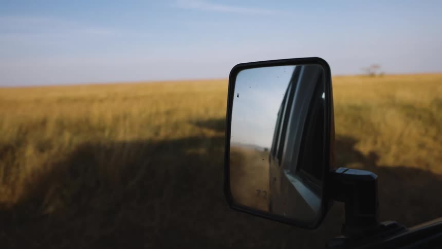 A vehicles side mirror reflects the open road behind with a vast field and clear sky