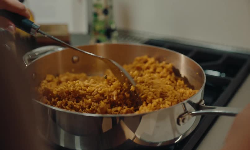 A closeup of a stainless steel pan on a stove with yellow rice being stirred by a persons hand using a spoon