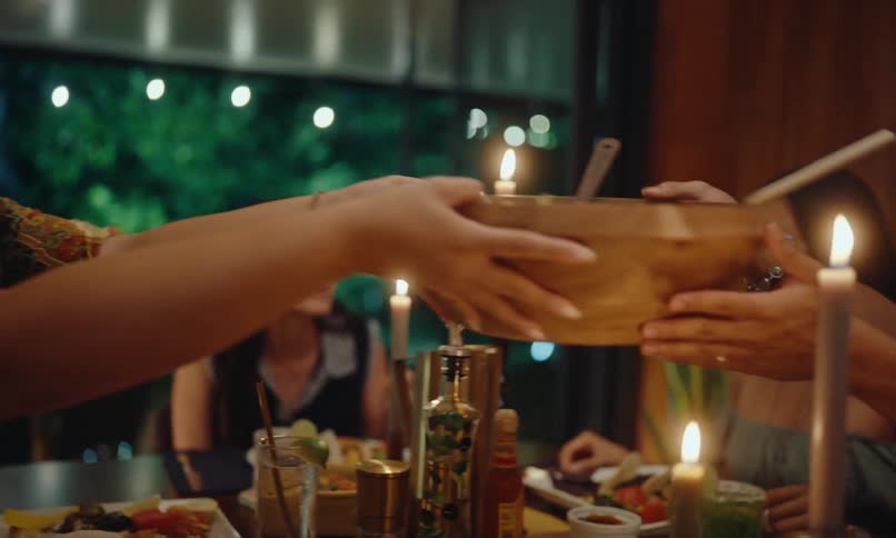 Some people are sitting around a table full of food and some of them are talking and one person hands a tray to another