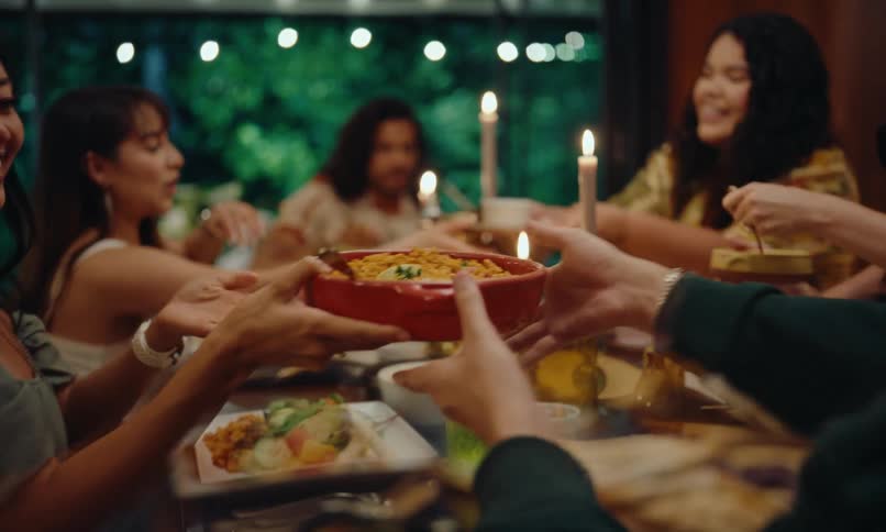 Several people around a table full of food and one of them is reaching a bowl of food for another person