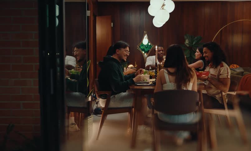 A group of some young and happy friends around a table are eating and behind them there is a dark wooden wall and above it a strong white lamp