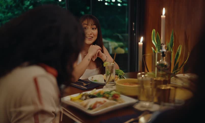 A young woman is talking to someone who is sitting at the same table as her and she is happy and on the table there are several plates of food