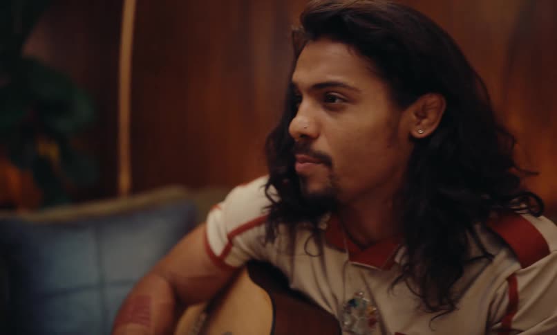 A young man with long hair wearing a retrostyle shirt engaged in conversation while holding a guitar