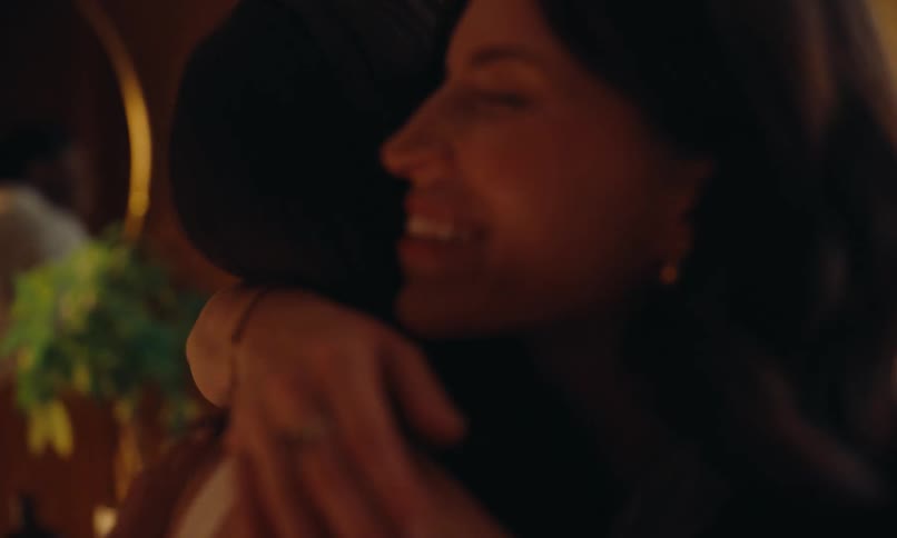 A woman and a man enter an apartment greeting their friends with hugs and handshakes while smiling