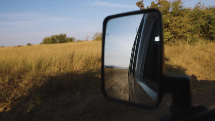 A cars side mirror reflects the vehicle on a dirt road surrounded by tall dry grass under a clear sky