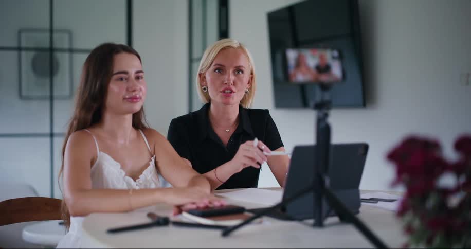 Two young women sitting at a table talking in front of an iphone filming in an indoor room