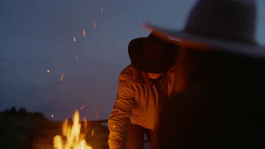 A man in a cowboy hat placing a piece of wood on a fire with a strong orange flame